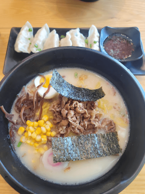 A bowl of ramen with meat, corn, seaweed, and toppings, alongside a plate of dumplings and dipping sauce.